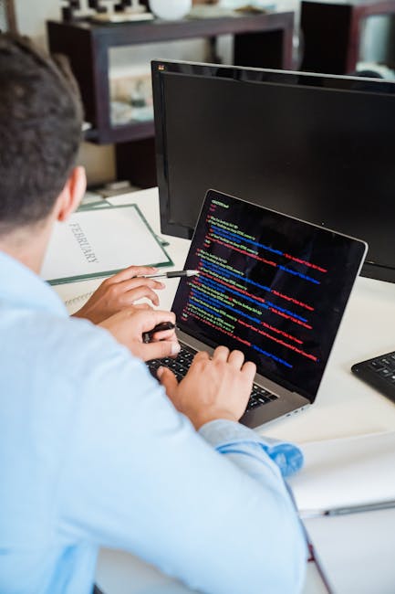 A person in a light blue shirt working on a laptop with code displayed on the screen, situated on a white desk. The desk also holds a closed green folder, a black monitor, and a remote control. In the