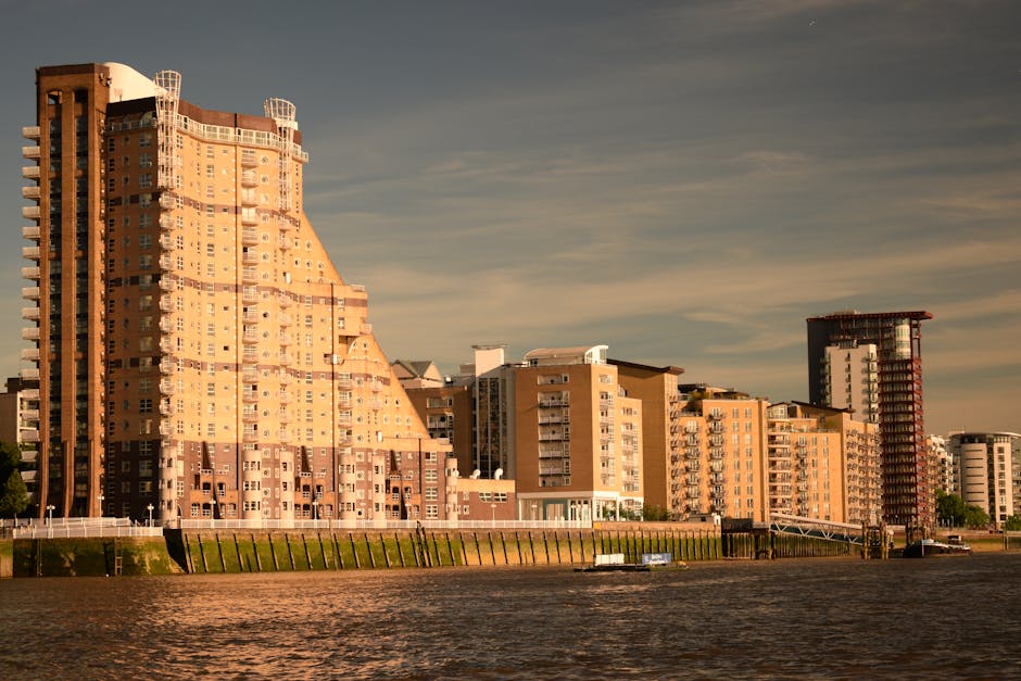 A daytime exterior view of two residential high-rise buildings, one modern with blue and white panels and large windows, the other a beige brick structure with multiple windows and balconies. Bright s